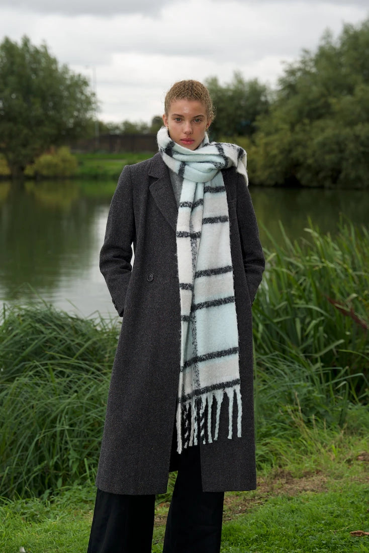 woman wearing extra long light blue, white and black plaid scarf with tassels in front of lake